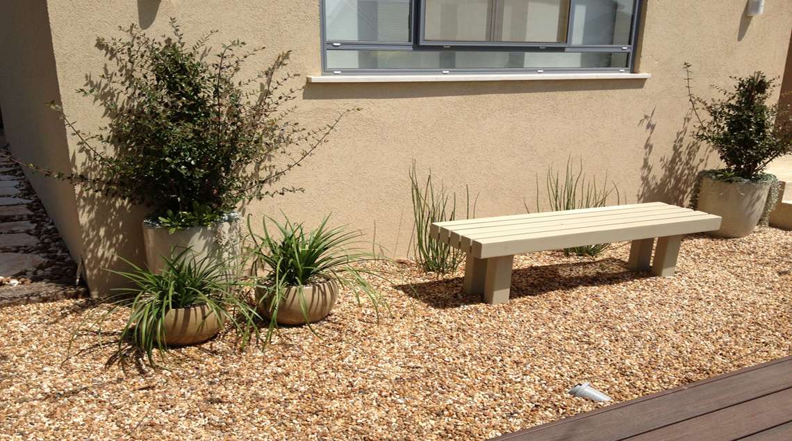 ספסל עץ בצבע שמנת מתחת לחלון המטבח. על מצע חלוקים קטנים בגוון חום. מאחוריו ובצידו צמחים ירוקים.