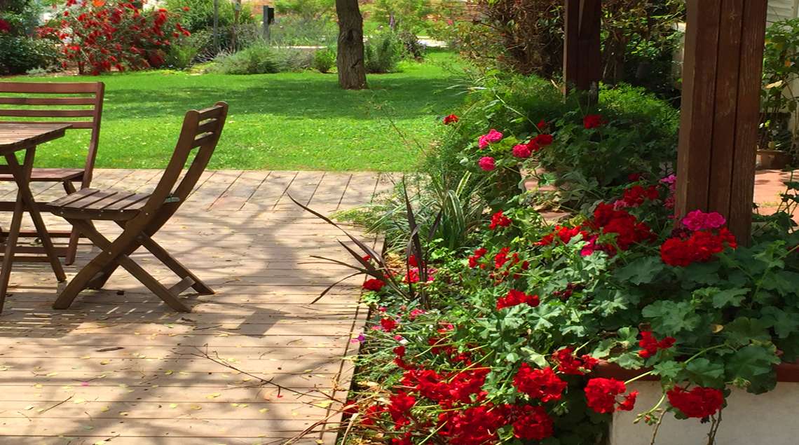 שולחן וכסאות עץ על דק ליד ערוגת גרניום פורחת באדום.