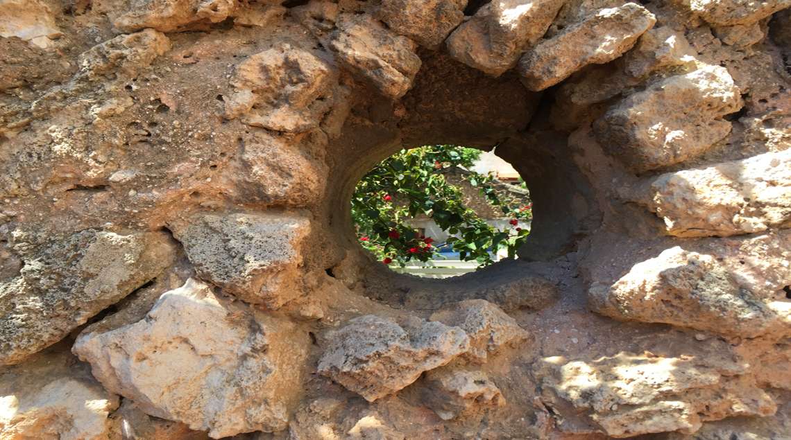 מבט לתוך חור עגול בקיר כורכר עתיק מחוץ לגינה פנימה.