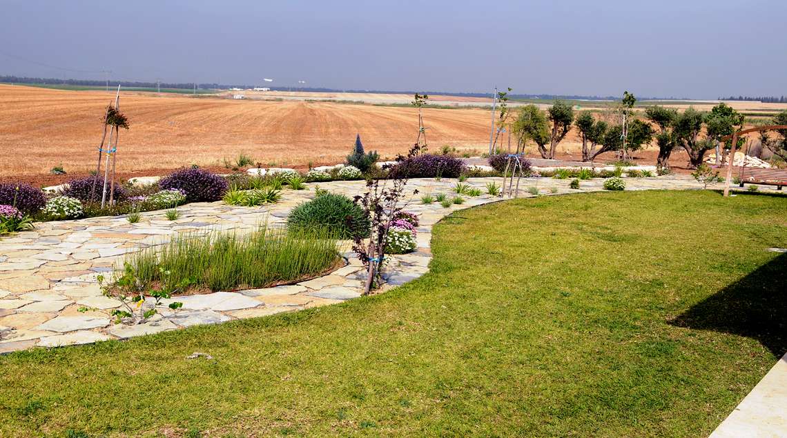 קו מפותל של מדשאה גובל בשביל אבנים ובתוכו ערוגות ואחריהם השדות