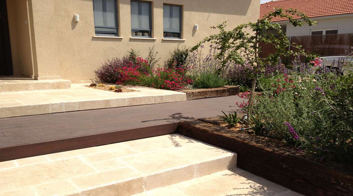 דק עץ ומדרגות אבן וערוגות פורחות באדום וסגול.