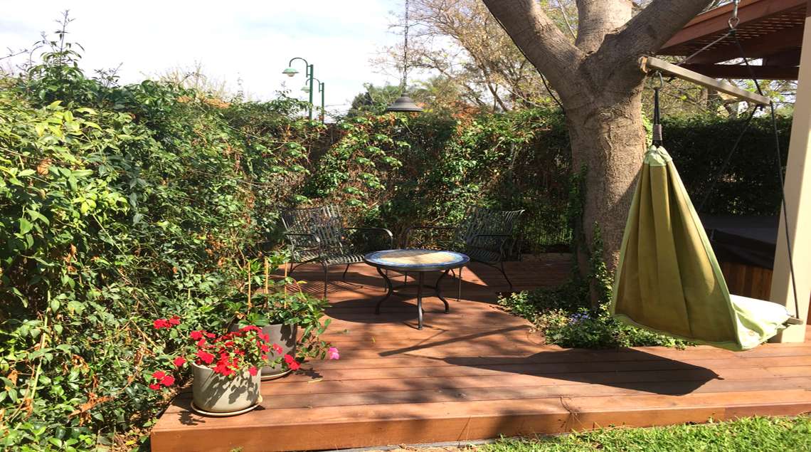 פינת ישיבה מדק, סביב עץ גדול. שולחן פסיפס קטן, שני כסאות וערסל ישיבה.