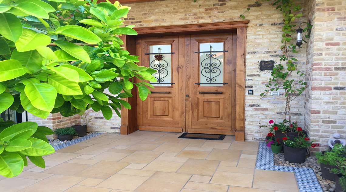 פאטיו הכניסה לבית. דלתות עץ גדולות, תיחום אריחים מצויירים של ערוגות סביב הבית ובהן שתילה בעציצים.