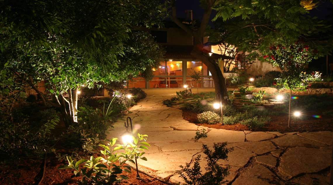 תאורת גינה בלילה משני צידיו של שביל אבני מדרך עם ערוגות.