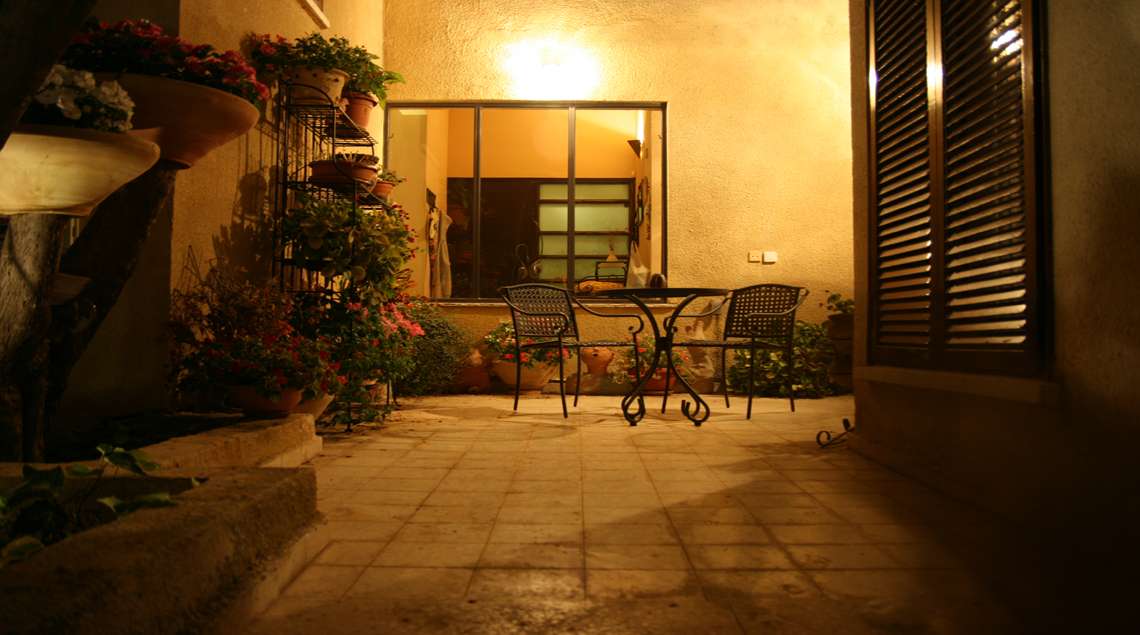פאטיו עם שולחן קטן ושני כסאות ועציצים סביב מואר בתאורת לילה.