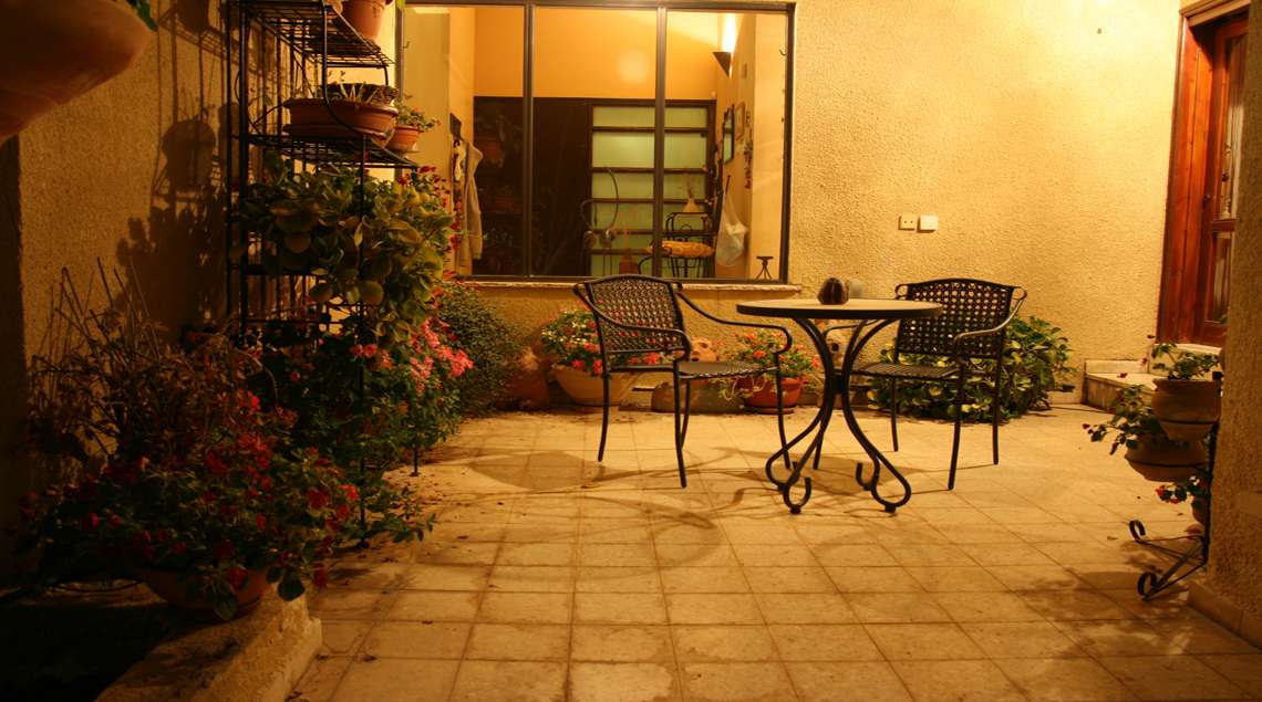 פאטיו עם שולחן קטן ושני כסאות ועציצים סביב מואר בתאורת לילה.