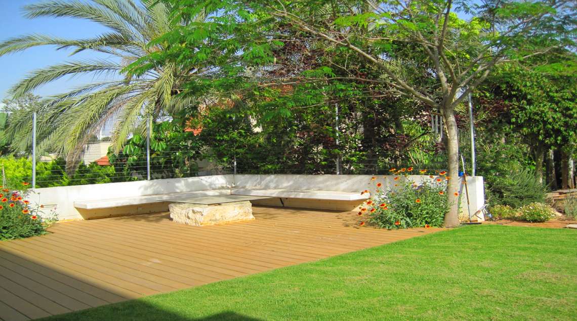 מדשאה, אחריה דק ועליו ספסל בטון לבן. מסביב עצים.