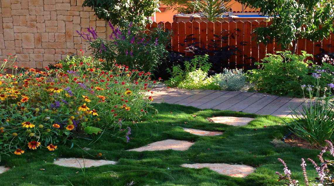 אבני מדרל מסודרות בתוך דשא גבעות ליד שביל דק עם ערוגוץ פרחים בגוונים של כתום סגול ירוק ואפור