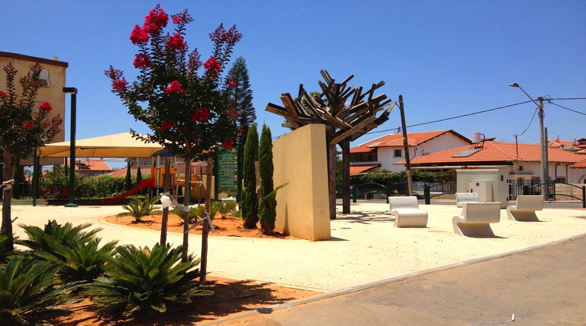 מבט לעבר קיר ההנצחה והפסל של שני העצים מברזל בצבע חלדה. ליד הפסל כסאות עגולים מבטון.